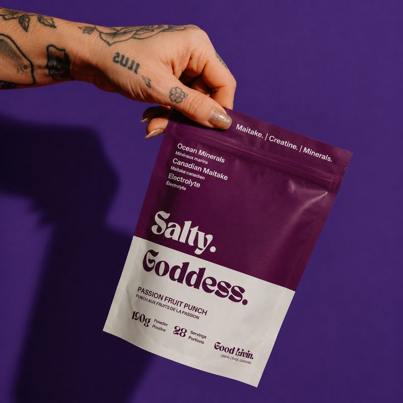 A tattooed hand holding a bag of Salty Goddess against a purple background